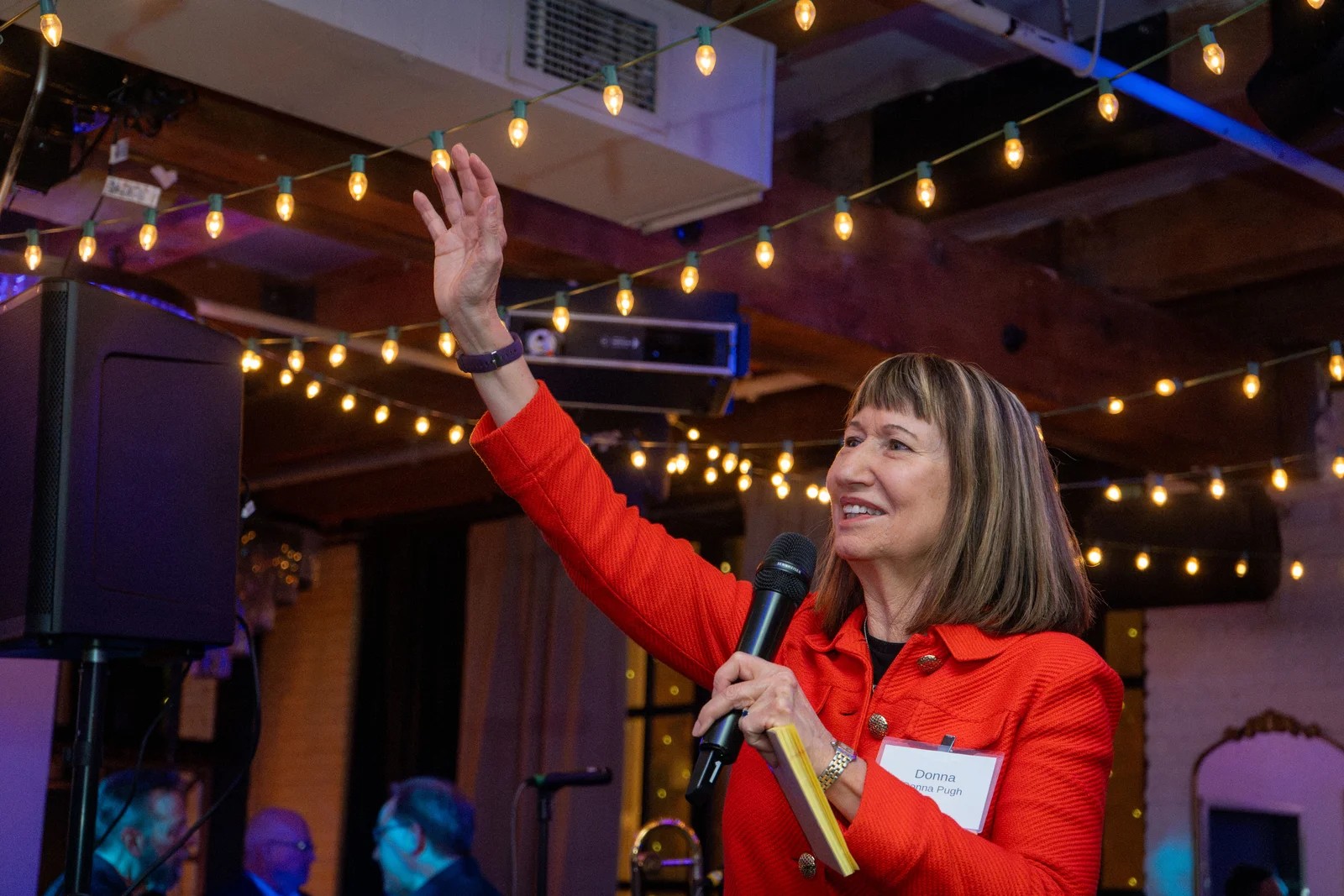 A host waving to the room, microphone in hand, under string lights at a Hubbard Inn celebration