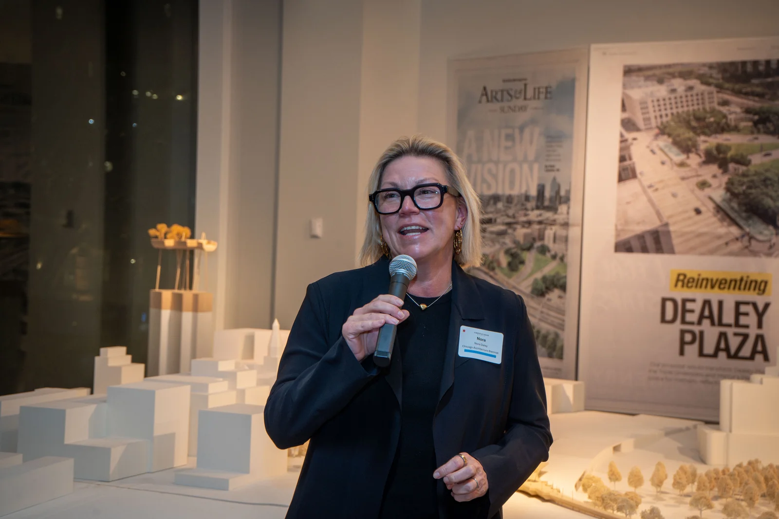A speaker with a microphone beside architecture models at the Magnificent Mile Association's Architecture Biennial event