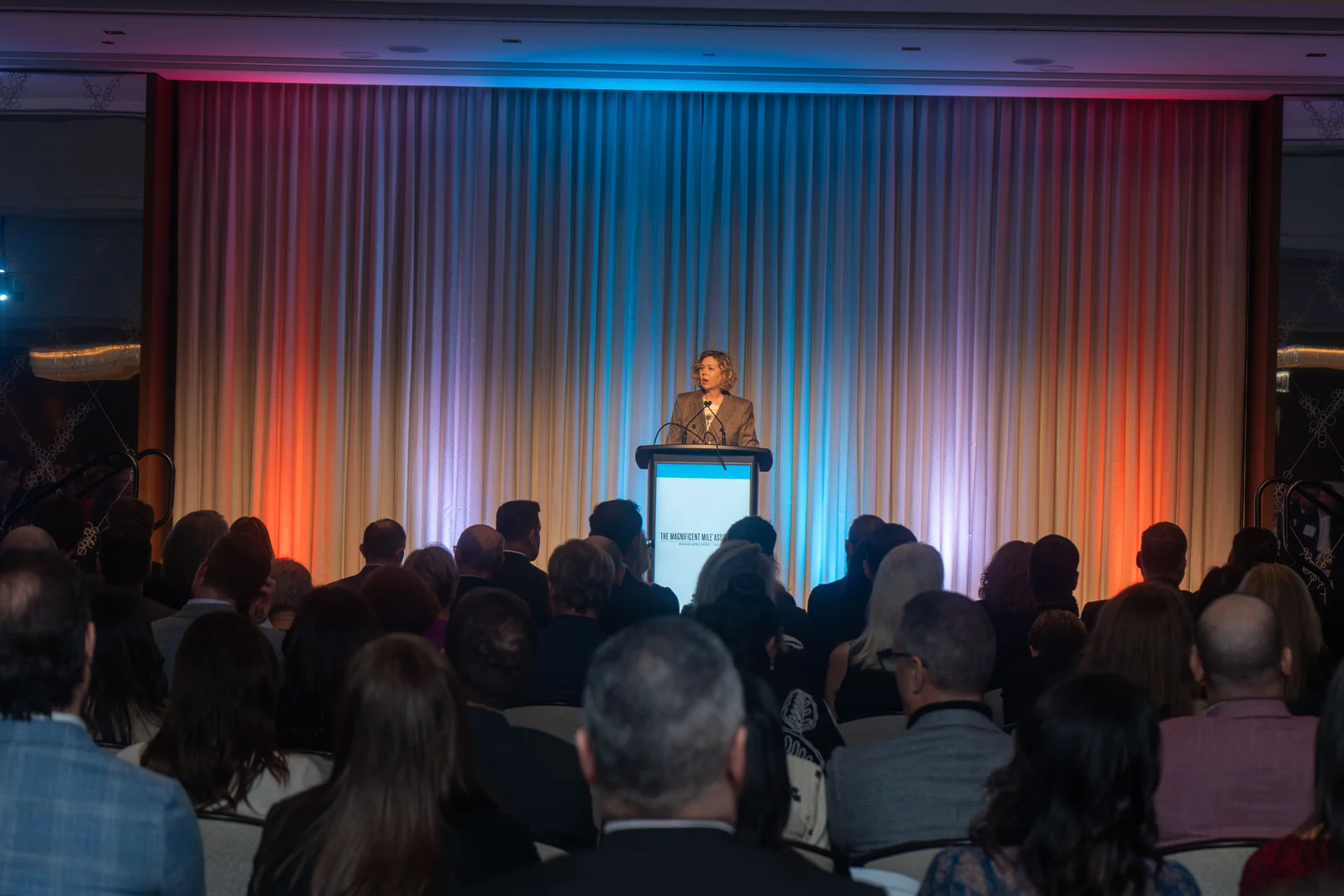Keynote speaker at the podium of a Magnificent Mile Association gala, the ballroom lit red and blue
